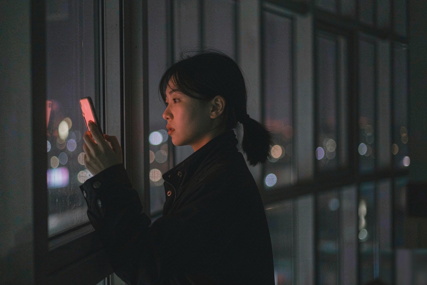 夜晚站在窗边看着城市灯火使用手机的人